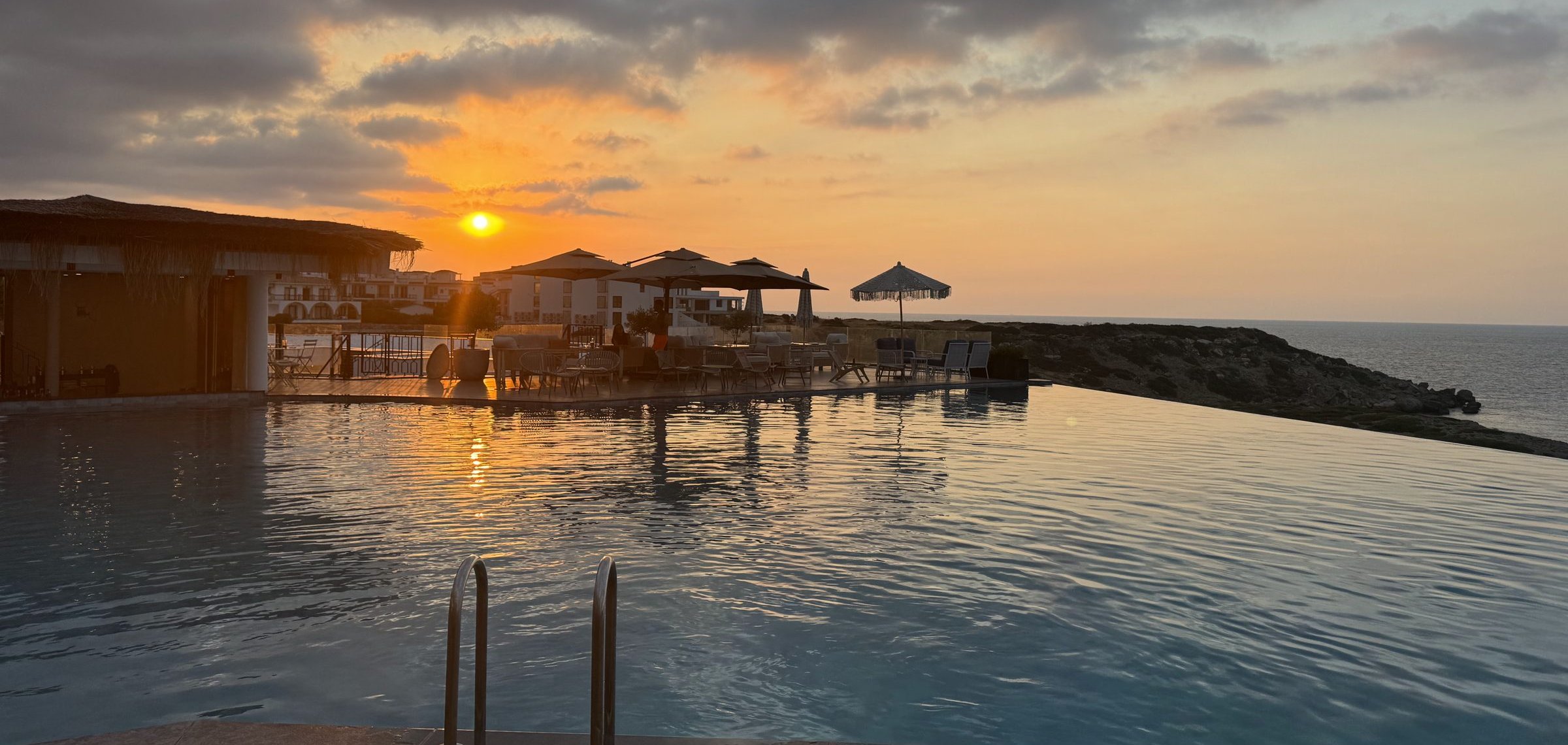 Infinity-Pool bei Sonnenuntergang mit Sonnenschirmen, Felsküste und Meer